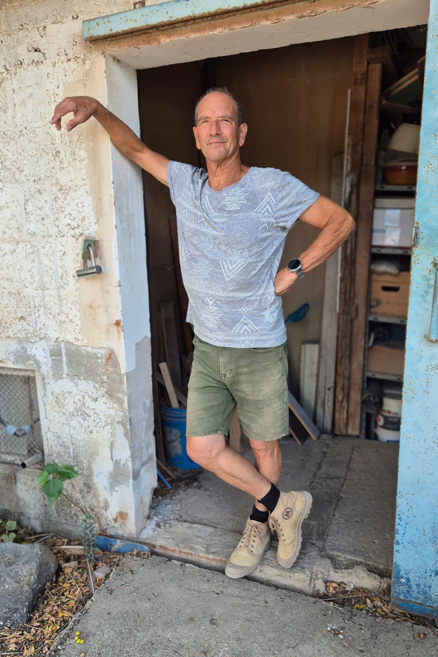Joseph Palmor standing in the open doorway of his studio, one arm resting on the frame, shelves and stacked timber behind him.